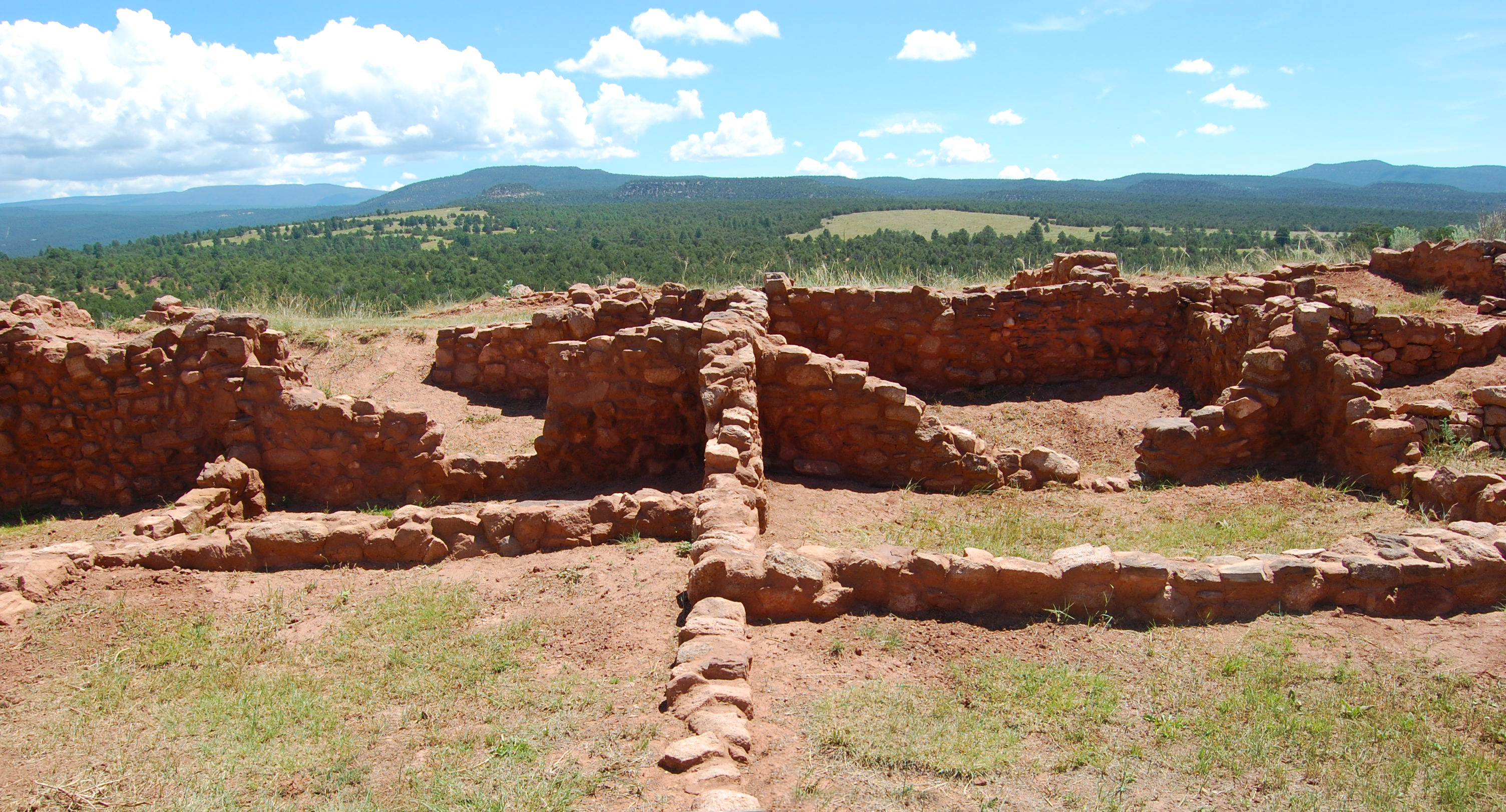 Pecos National Historic Site - Ruins of Pecos Pueblo; The foreground contains the remnants of Puebloan structures. On the ground, there is an intersection of earthen bricks, creating four quadrants. Behind this, are 3-4" high walls, which sit along the intersecting grid. The ruins sit atop a hill overlooking a large valley vista and partly cloudy sky.
